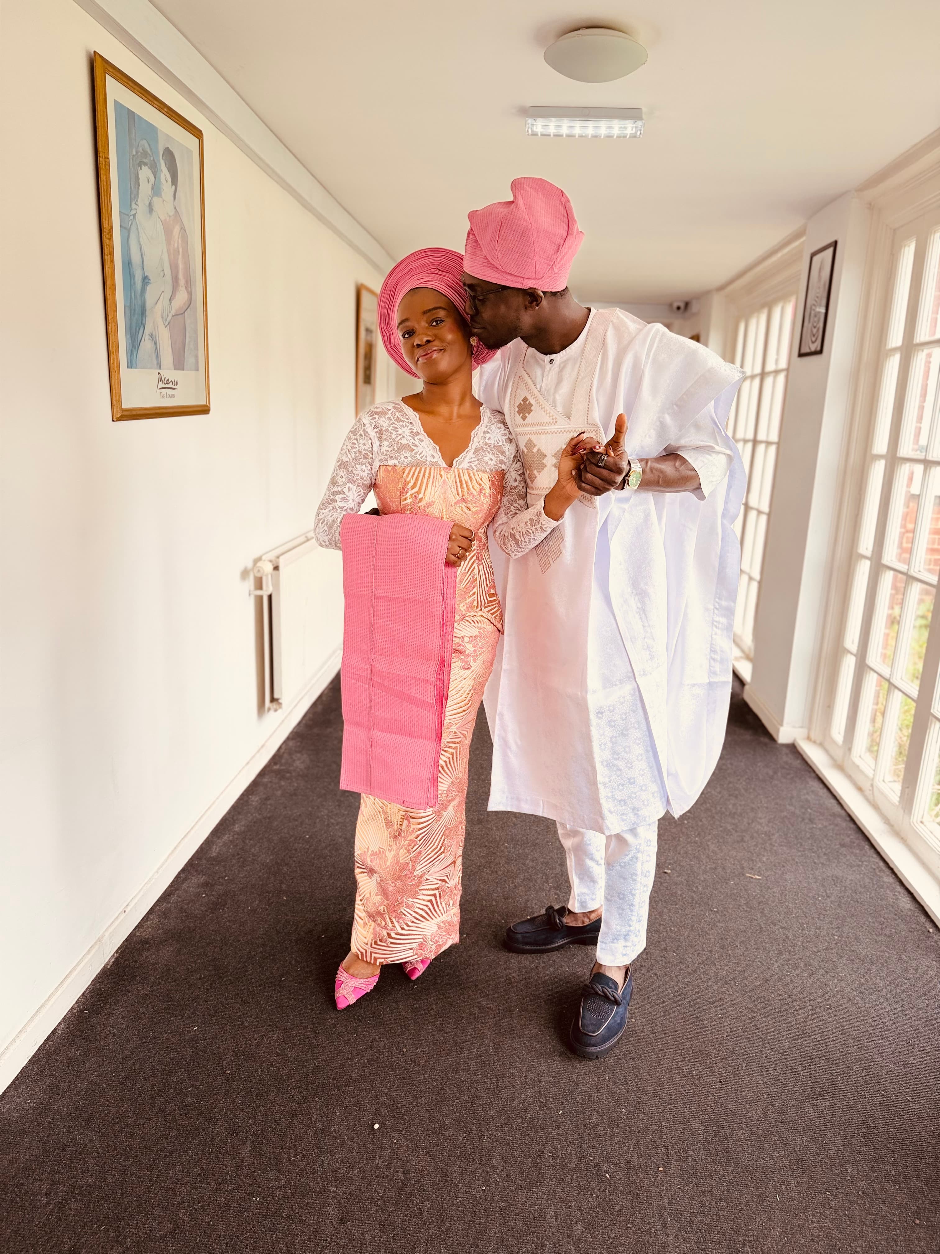 Ayo and Ola dressed in traditional aso-oke, holding hands in a hallway